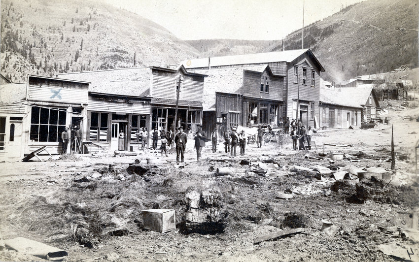 The morning after the 1899 fire in Gilman, which destroyed approximately half of the town. The Fleck Clothing Store is at the left of the photo and is marked with an "X". People standing survey the damage.