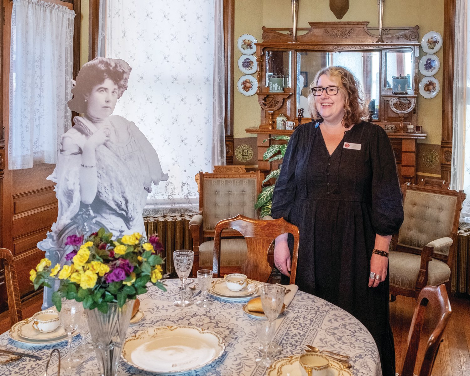 Andrea Malcomb, director of the Molly Brown House Museum, exchanges glances with a cardboard cutout of Margaret Tobin Brown at the Brown family dinner table. The bowls at each setting suggest that the family&rsquo;s first course might have been consomm&eacute;, a clear broth often served around the turn-of-the-century.