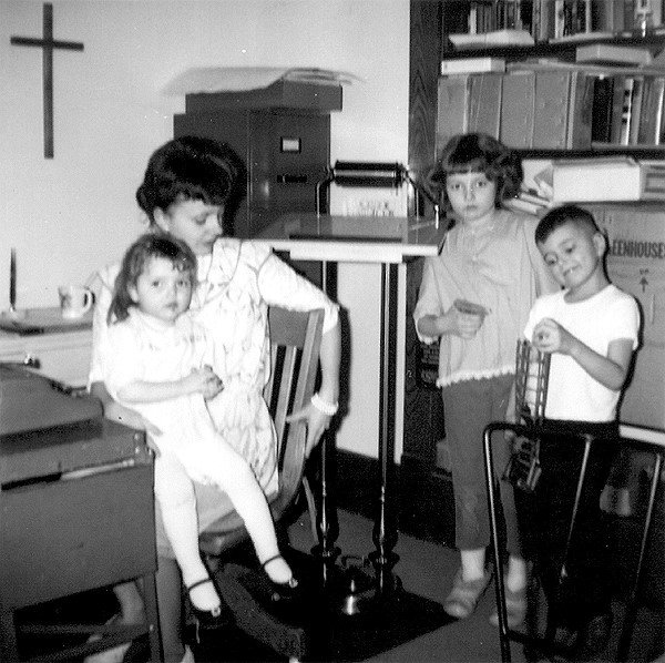 Since age 10, Katie's lifelong passion was to serve God and His Church. A dutiful pastor’s wife and mother, she is shown readying the church bulletin. Photographed In the pastor’s church study at Avoca, Nebraska, she is surrounded by her two daughters and oldest son, Darrell. Katie, now 88, constantly looks for things she can do at her Aledo church, still trying to inspire others. 