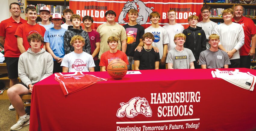 Harrisburg senior Trace Combs (seated in black shirt) signed a letter of intent to play basketball next season at NCAA DII Quincy University. Head coach Kyle Fisher (standing left) and father and assistant coach Steve Combs (standing right) along with the Harrisburg Bulldogs basketball team cheered on their senior scoring at the signing event.