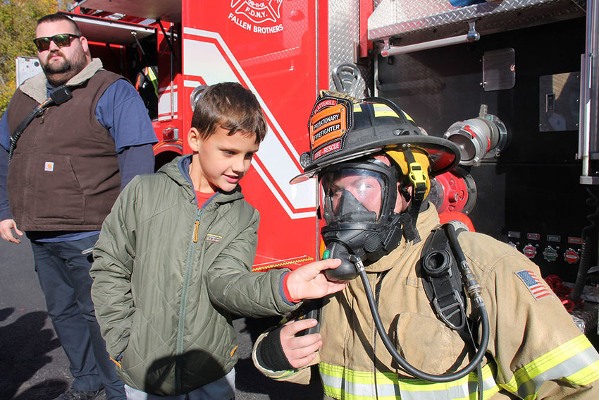 Leptondale Elementary School Grade 2 student Max Owen checks out a self-contained breathing apparatus (SCBA) worn by Plattekill Fire Department member Nick Platoni) as Cronomer Valley Fire Department First Lieutenant Max Villalonga looks on .