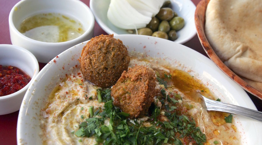 A bowl of hummous is served with olive oil, falafel balls and sprinkled with chopped parsley alongside fresh-baked pita bread, olives, a fresh onion, red chili paste and chili and lemon juice in a restaurant in Tel Aviv.