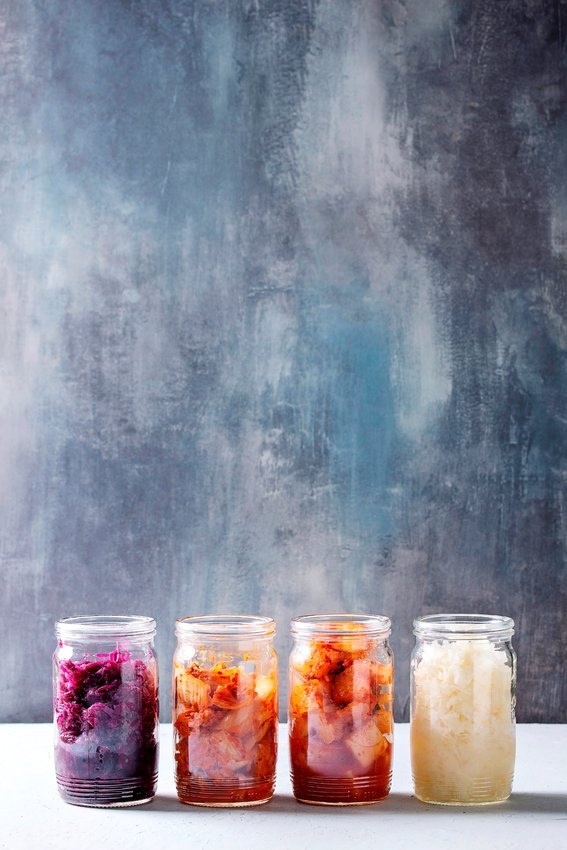 Variety of fermented food korean traditional kimchi cabbage and radish salad, white and red sauerkraut in glass jars in row over grey blue table.