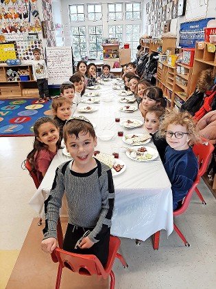 HALB Lev Chana students are seated in front of seder plates, posing happily for a photographer before their model seders got underway.