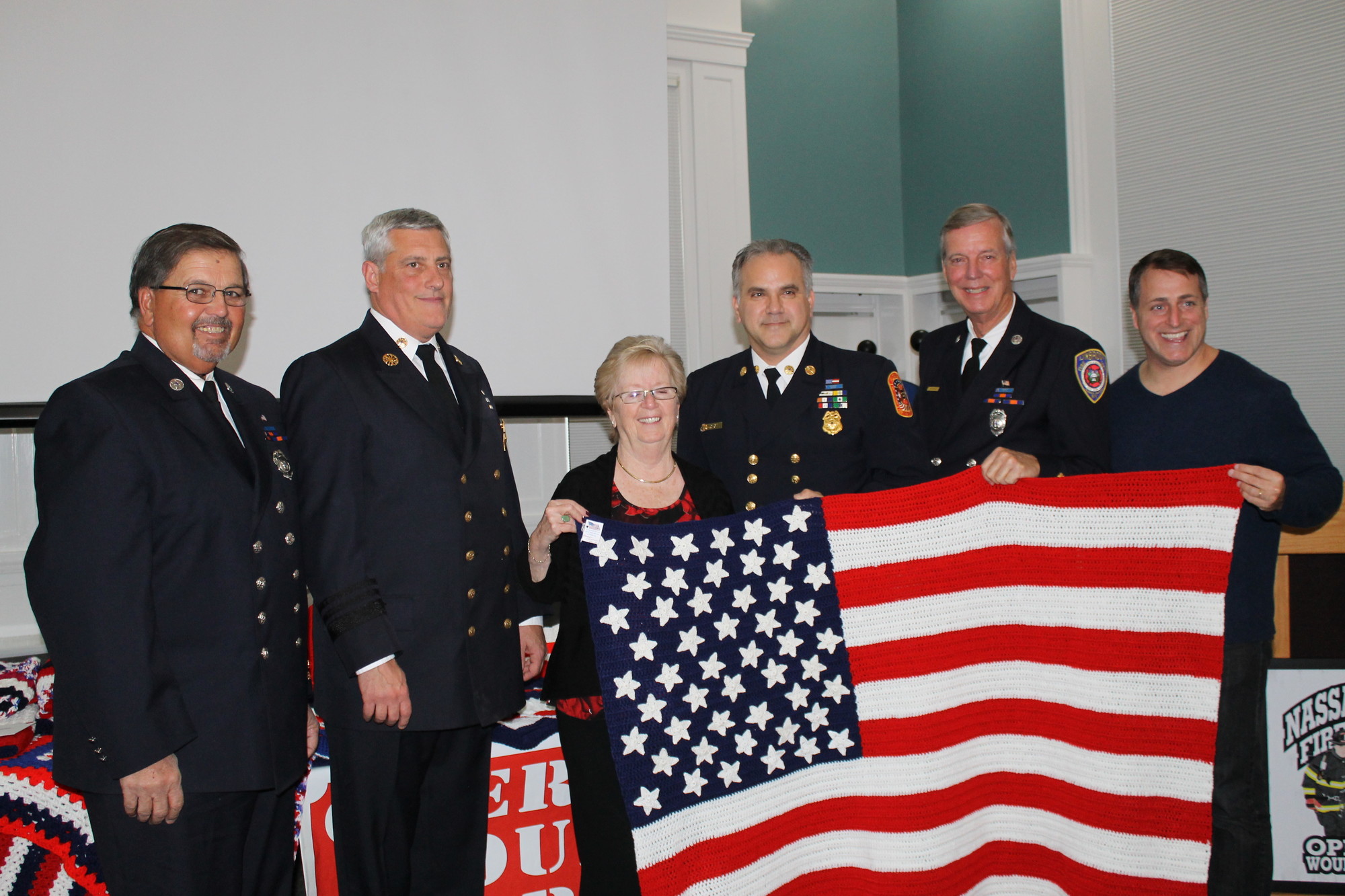 Lynbrook resident Barbara Barnhart, president and founder of Loops of Love, held one of the American flag blankets sewn by one of its members. Joining her were, from left:  Richard Straub, Lynbrook Fire Department; Bill Graham, Garden City Fire Department; Joe O'Grady, Floral Park Fire Departmentand Chairman of NCFF-OWW; Steve Grogan, Lynbrook Fire Department, and Assemblyman Brian Curran.
