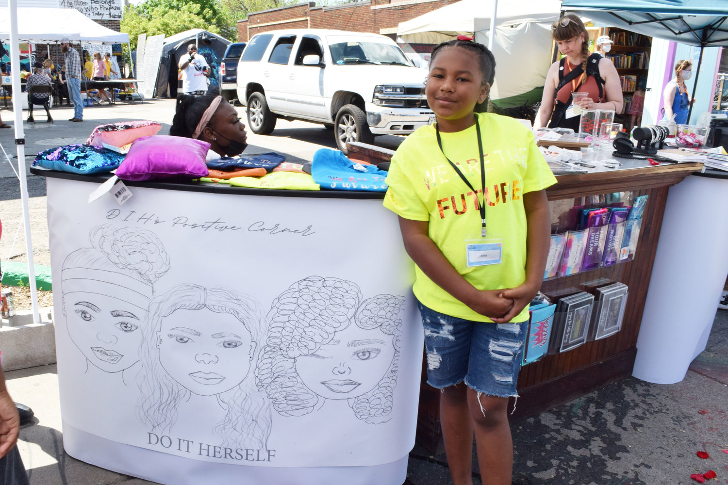 Wearing a yellow t-shirt that reads “We are the Future,” Innocynce stands at her booth, D.I.H. (Do It Herself) Positive Corner. Positive Corner is a pop-up boutique shop run by youth entrepreneurs teaching other youth how to not become juvenile delinquents. The drawing with the shop name, by Heaven Rain, features from left to right: Deztiny, Innocynce and Heaven. At 11 years old, Innocynce also opened the evening concert singing the Black National Anthem.