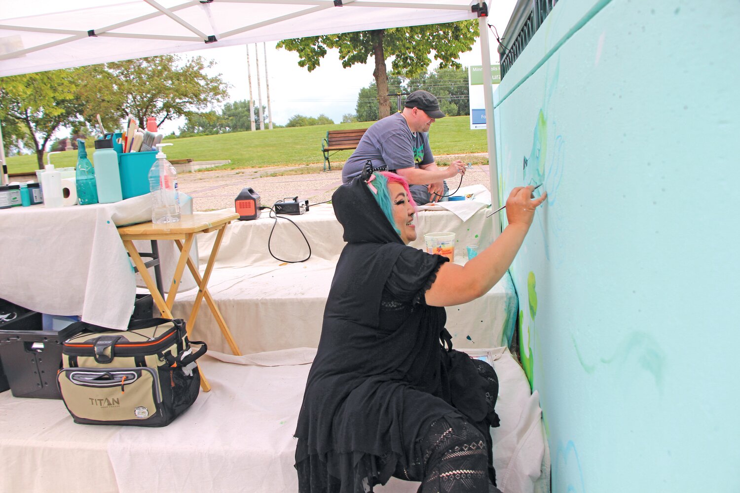Artists Kao Lee Thao and Ash Kubesh work on a mural at Boom Island Park that fills two sides of a concrete wall near the river. Thao painted a woman on one side of the mural wall. “Over there she symbolizes mother nature and earth,” Thao said. “When you see a woman in my pieces, it’s like she’s a storyteller. Like she’s telling a story about the importance of water and parks and nature.” She described how in Hmong culture, women often hand stitch their clothing and costumes. The woman Thao painted in bright colors on the mural, is inspired by and in honor of Hmong women.