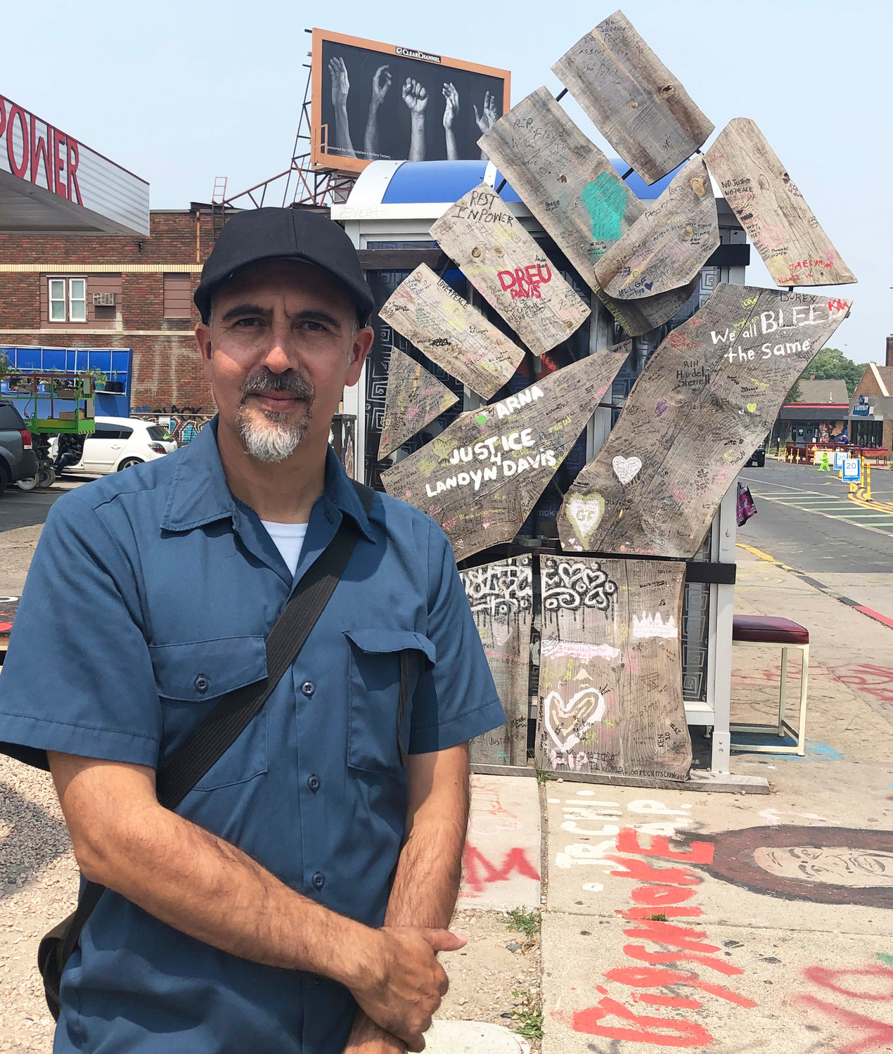 Xavier Tavera at a wooden fist sculpture at George Floyd Square, his billboard “Unified” visible in the background. (Photo by Jill Boogren)
