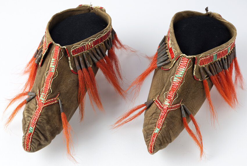 This “Great Lakes First Nation Pair of Moccasins,” circa 1770-80, are black-dyed deerskin sewn with sinew, decorated with porcupine quillwork and fringed with red dyed deer hair tassels inserted in tinned iron cones.
