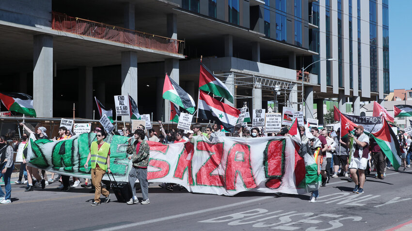 A protestor in a camouflage jacket leads the protest down Fillmore Street on March 30, passing the Arizona State University Health North building. Behind him, a “SAVE GAZA” banner and Syrian-Palestinian flags are waved. Chants heard at this point included “Rafah will be liberated. Free, free Palestine”and “ASU! ASU! Shame on you!  Shame on you!”