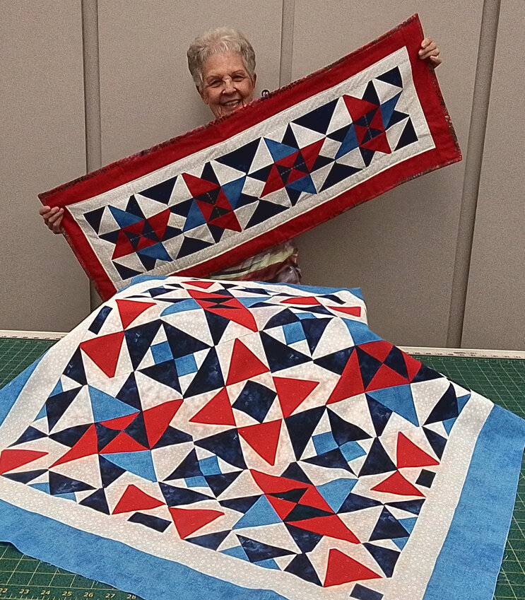 Marilyn Zenz chose shades of red, off-white and blues for her table runner, which she holds, and table topper. The topper is sewn together on-point with setting blocks that mimic the layout of the larger quilt.