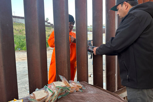 Kedian William, 38, of Jamaica, hands a phone charger to volunteer Pedro Rios while waiting between two border walls in San Diego for agents on Friday, March 29, 2024.