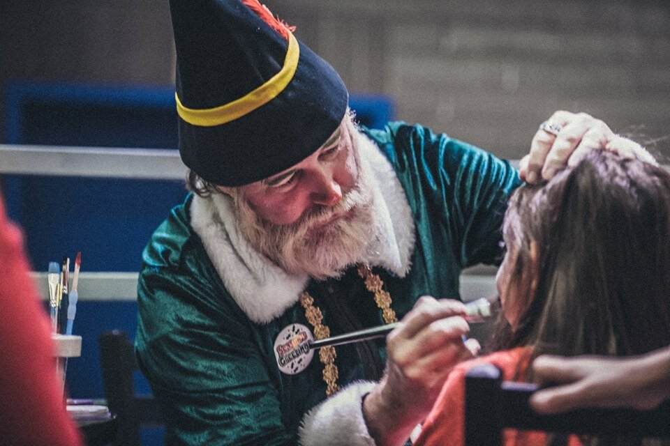 An actor dressed as an elf does face-painting for a child.